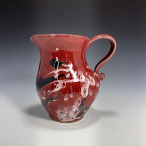 Op de afbeelding: Handgemaakte keramische kan met een rode glazuur, met zwarte en witte abstracte ontwerpen. De kan heeft een gebogen handvat en een schenktuit. Deze decoratieve kan is geschikt voor het serveren van dranken.