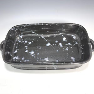 Puede incluir: Una fuente rectangular para hornear de cerámica negra con esquinas redondeadas y dos asas pequeñas. La fuente tiene un patrón blanco moteado en la superficie. La fuente para hornear está sobre un fondo blanco.