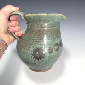 Op de afbeelding: Handgemaakte keramische kan met een groene en bruine glazuur. De kan heeft een handvat, een tuit en decoratieve bloemen- en cirkelontwerpen. Het aardewerk heeft een rustieke, getextureerde uitstraling.