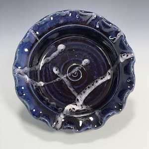 May include: A handmade ceramic pie plate with a dark blue glaze and a ruffled edge. The interior features a spiral design with white accents. The plate is approximately 9 inches in diameter, perfect for serving pies or other baked goods.