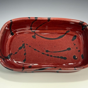 Puede incluir: Una fuente rectangular de cerámica para hornear con un esmalte rojo brillante y un diseño de salpicaduras negras. La fuente tiene esquinas redondeadas y asas pequeñas en cada extremo. Una pieza decorativa para la cocina.
