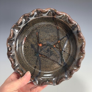 May include: A handmade ceramic pie plate with a speckled brown interior and a ruffled edge. The plate features a dark brown rim and is decorated with abstract lines in orange, blue, and black.