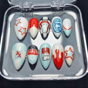 Puede incluir: Un juego de diez uñas postizas de forma ovalada en una caja de plástico transparente. Las uñas están pintadas con varios diseños, incluyendo estrellas, labios y la palabra "Lost". Los colores incluyen blanco, rojo, negro y azul.