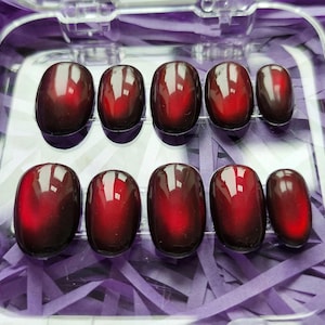 May include: A set of ten oval-shaped, glossy, dark red press-on nails. The nails are arranged in two rows inside a clear plastic container, with a purple paper background. The nails have a gradient effect, transitioning from dark red to a lighter shade.