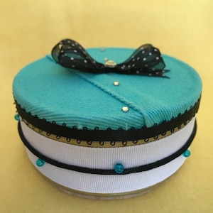 May include: A round turquoise box with a black and white polka dot bow on top. The box is decorated with black lace and white ribbon, and has turquoise beads around the bottom.