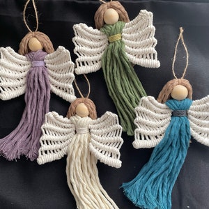 May include: Four handmade macrame angel ornaments with wooden heads and different colored yarn tassels. The angels have white wings and are hanging from a string.