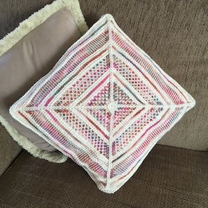 Könnte beinhalten: Ein gehäkeltes Kissen mit einem geometrischen Muster in Rosa-, Weiß- und Brauntönen. Das Kissen ist mit einer zarten, durchbrochenen Masche gearbeitet.