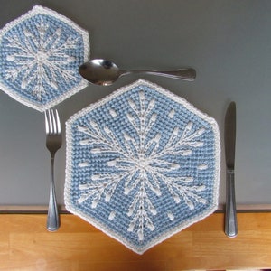 Pode incluir: Dois tapetes de crochê em forma de floco de neve em azul e branco. O tapete maior está no centro da imagem, e o menor está à esquerda. Um garfo e uma colher estão colocados na mesa.
