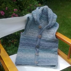 Peut inclure: Un gilet en tricot rayé bleu et blanc avec une capuche et des boutons en bois. Le gilet est posé sur une chaise blanche.