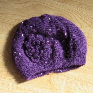 Puede incluir: Boina de punto morado con una gran flor y lentejuelas.