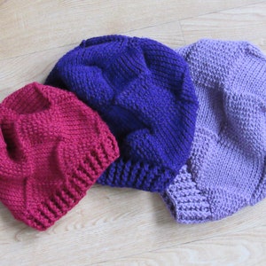 May include: Three knitted hats in different colors: a deep purple, a bright pink, and a light lavender. The hats are all made with a textured stitch and have a slouchy, relaxed fit.