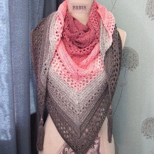 Op de afbeelding: Een gehaakte sjaal met een roze, grijs en wit ombre-patroon. De sjaal heeft een delicaat kantpatroon en is afgewerkt met kwastjes. De sjaal wordt getoond op een mannequin.