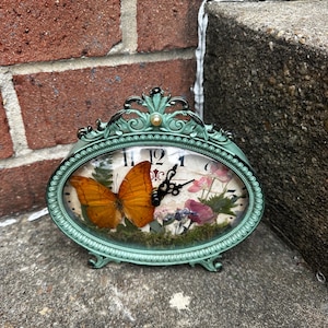 May include: An oval-shaped table clock with a distressed teal metal frame. The clock face features a large orange butterfly, green moss, and pink floral accents behind the glass. Black clock hands are centered over the butterfly.