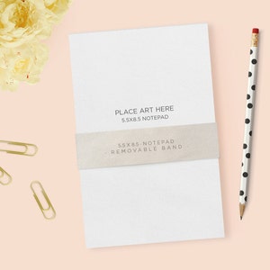 May include: A white notepad with a removable band, text that reads "PLACE ART HERE 5.5X8.5 NOTEPAD" and "5.5X8.5 NOTEPAD - REMOVABLE BAND". A black and white polka dot pencil and gold paper clips are also present. The background is a light pink.