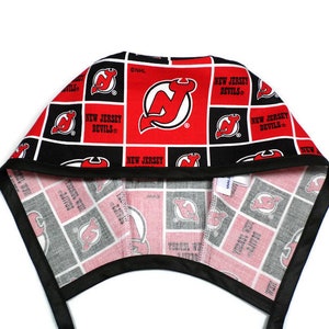 May include: A black and red fabric scrub hat with the New Jersey Devils logo repeated throughout. The hat has a black elastic band and ties.