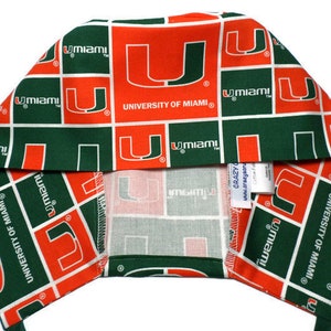 Puede incluir: Un turbante de tela verde y naranja con el logotipo de la Universidad de Miami repetido en toda la superficie. El logotipo es una 'U' blanca dentro de un cuadrado naranja con las palabras 'University of Miami' en texto blanco.