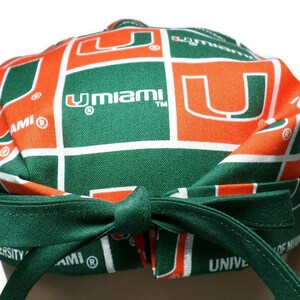 Puede incluir: Bandana verde y naranja con el logotipo de la Universidad de Miami y el texto "UMiami" repetido en toda la superficie.