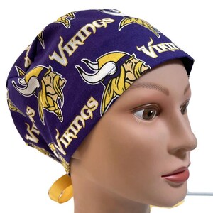 May include: A purple and gold Vikings print scrub cap. The cap features the team's logo and the word "Vikings" in gold. The cap has a yellow tie in the back.