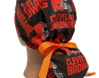 May include: A black and brown scrub cap featuring the words "Cleveland Browns" and football helmets in red. The cap is tied with an orange ribbon. The cap is designed to be worn on the head.