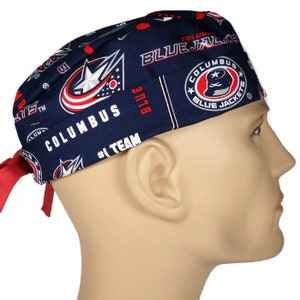May include: Blue and red surgical scrub hat with a repeating pattern of the Columbus Blue Jackets logo. The hat has a tie on the back.