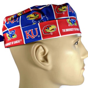 May include: A blue, red, and white patterned scrub cap featuring the University of Kansas Jayhawks logo and text. The cap is made of fabric and has a tie in the back. The text "KU" and "THE UNIVERSITY OF KANSAS" are visible.