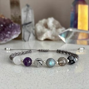 May include: A handmade bracelet featuring a series of colorful gemstone beads, including white, purple, and gray, strung on a gray cord. The beads are interspersed with small silver-colored beads. The bracelet is displayed on a white surface.