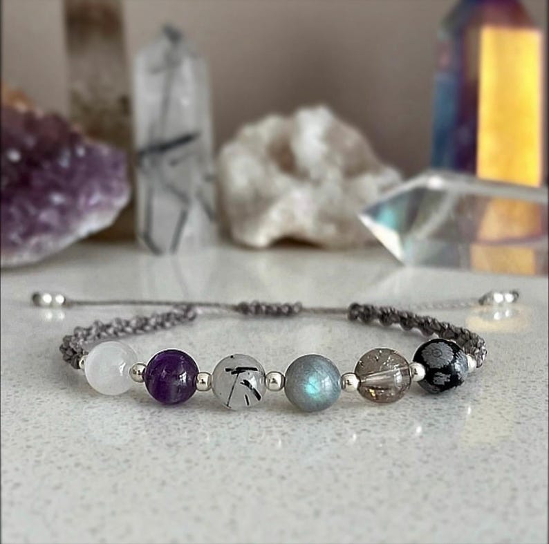 May include: A handmade bracelet featuring a series of colorful gemstone beads, including white, purple, and gray, strung on a gray cord. The beads are interspersed with small silver-colored beads. The bracelet is displayed on a white surface.