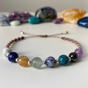 May include: A brown cord bracelet with seven gemstone beads. The beads are white, blue, yellow, green, blue, blue-green, and purple.
