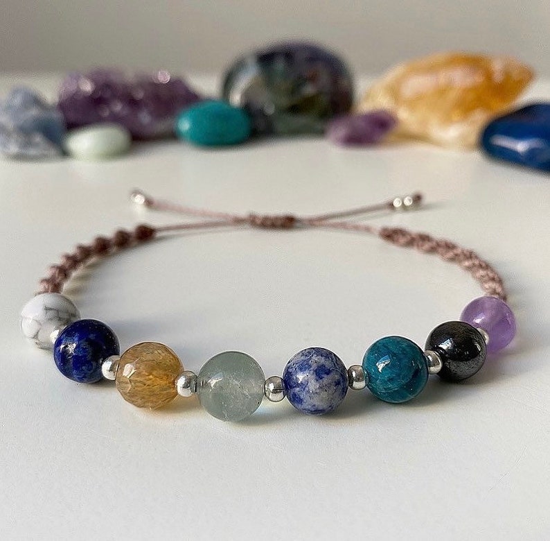 May include: A brown cord bracelet with seven gemstone beads. The beads are white, blue, yellow, green, blue, blue-green, and purple.