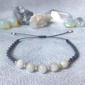 Puede incluir: Un brazalete de cuerda gris con cinco cuentas de piedra lunar blanca y cuentas espaciadoras plateadas.