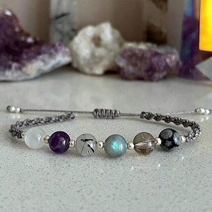 May include: A gray macrame bracelet featuring a series of gemstone beads. The beads include white, purple, clear with black inclusions, iridescent blue-green, brown, and black snowflake patterns. The bracelet has adjustable silver-toned end beads.