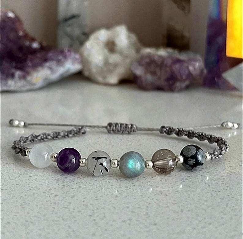 May include: A gray macrame bracelet featuring a series of gemstone beads. The beads include white, purple, clear with black inclusions, iridescent blue-green, brown, and black snowflake patterns. The bracelet has adjustable silver-toned end beads.
