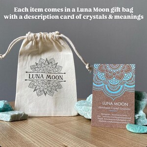 May include: A beige drawstring bag with the text "LUNA MOON" and a decorative floral design. A business card with the text "LUNA MOON Handmade Crystal Creations" is next to the bag. The image also includes crystals.