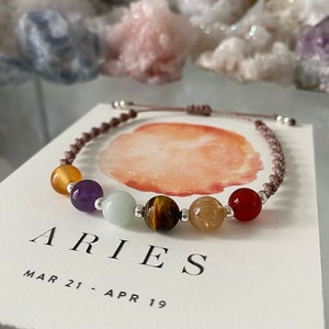 May include: A bracelet for the Aries zodiac sign, featuring a series of colourful gemstone beads, including yellow, purple, green, and red, strung on a brown cord. The bracelet is displayed on a card with the word "ARIES" and the dates "MAR 21 - APR 19".