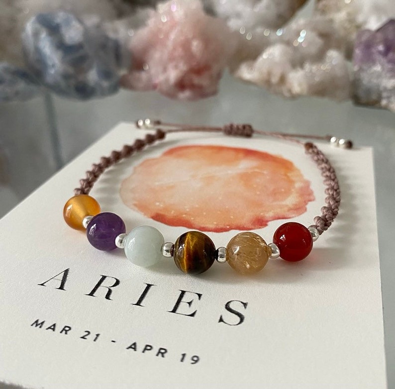 May include: A bracelet for the Aries zodiac sign, featuring a series of colourful gemstone beads, including yellow, purple, green, and red, strung on a brown cord. The bracelet is displayed on a card with the word "ARIES" and the dates "MAR 21 - APR 19".
