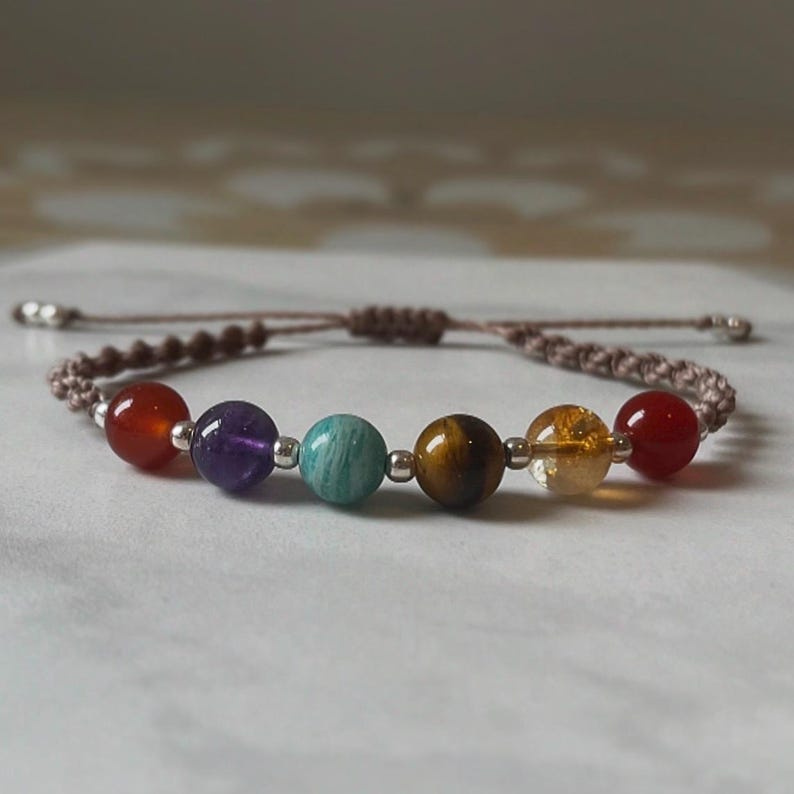 May include: A chakra bracelet with seven coloured gemstones: red, purple, teal, brown, yellow, and orange. The beads are strung on a brown cord with adjustable sliding knots. The bracelet is designed to represent the body's energy centres.
