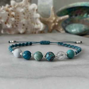May include: A teal macrame bracelet with six gemstone beads in white, teal, green, and blue. The adjustable bracelet has silver-coloured metal accents and is displayed on a marble surface. The bracelet is a piece of jewellery.
