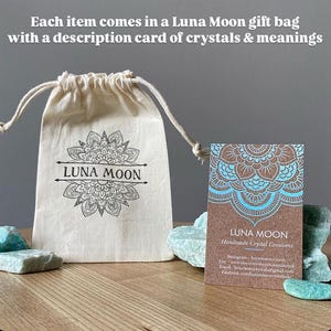 May include: A white cotton drawstring bag with the text "LUNA MOON" printed on it. The bag is sitting on a wooden surface next to a brown card with a blue mandala design and the text "LUNA MOON Handmade Crystal Creations".