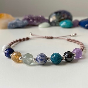 May include: A brown cord bracelet with seven gemstone beads. The beads are a mix of blue, green, purple, and brown.