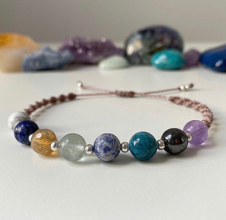 May include: A brown cord bracelet with seven gemstone beads. The beads are a mix of blue, green, purple, and brown.