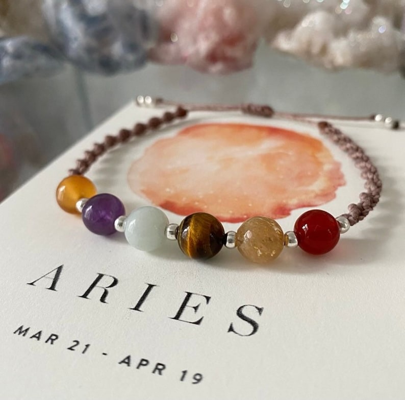 May include: A bracelet with seven gemstones in various colours, including yellow, purple, and red, strung on a brown cord. The bracelet is displayed on a card with the word "ARIES" and the dates "MAR 21 - APR 19".