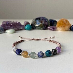 May include: A bracelet with a brown cord and seven gemstone beads. The beads are white, blue, yellow, green, blue, black, and purple.