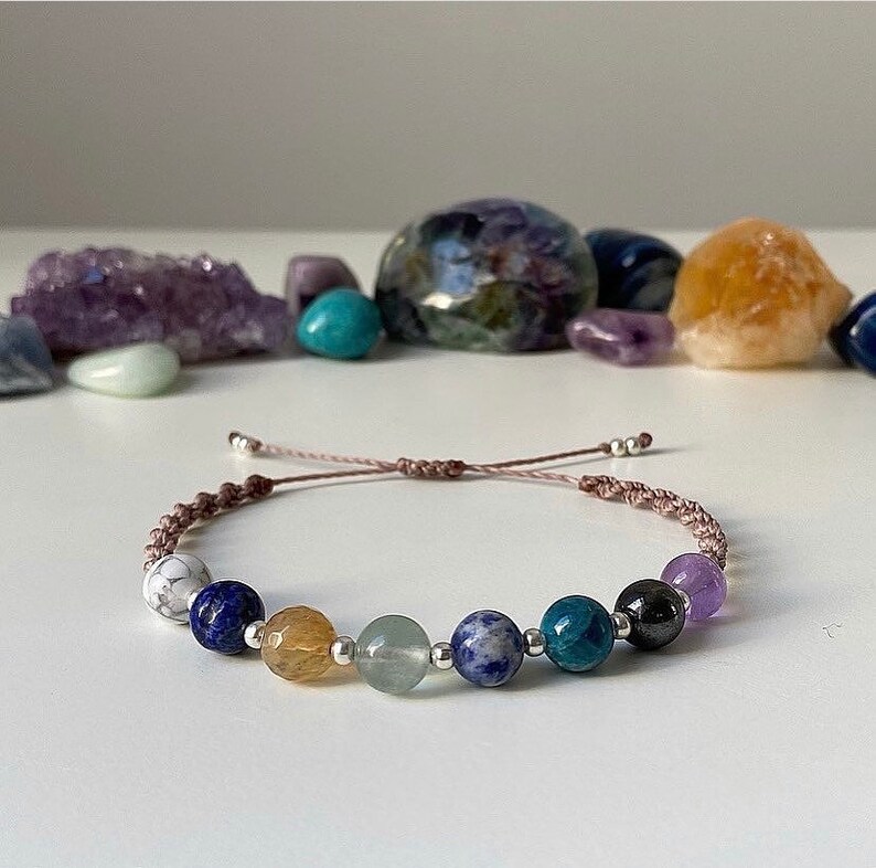 May include: A bracelet with a brown cord and seven gemstone beads. The beads are white, blue, yellow, green, blue, black, and purple.