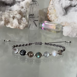 May include: A grey macrame bracelet with silver beads and moon charms. The bracelet features black snowflake obsidian, smoky quartz, labradorite, and moonstone beads.