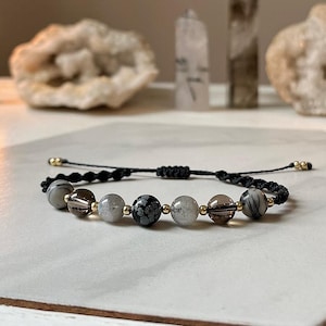 May include: A black cord bracelet with seven round, grey, white, and black gemstone beads. The beads are separated by gold spacers.