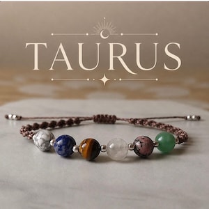 May include: A brown macrame bracelet with seven colorful gemstone beads. The bracelet features a white and gray speckled bead, a blue bead, a tiger's eye bead, a clear bead, a pink bead, and a green bead. The word "TAURUS" is displayed above the bracelet.