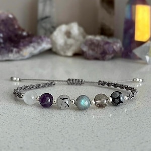 May include: A gray macrame bracelet featuring a series of gemstone beads in white, purple, black, and blue-green hues. The bracelet has silver-colored end beads and a sliding knot closure. The background includes blurred crystals.
