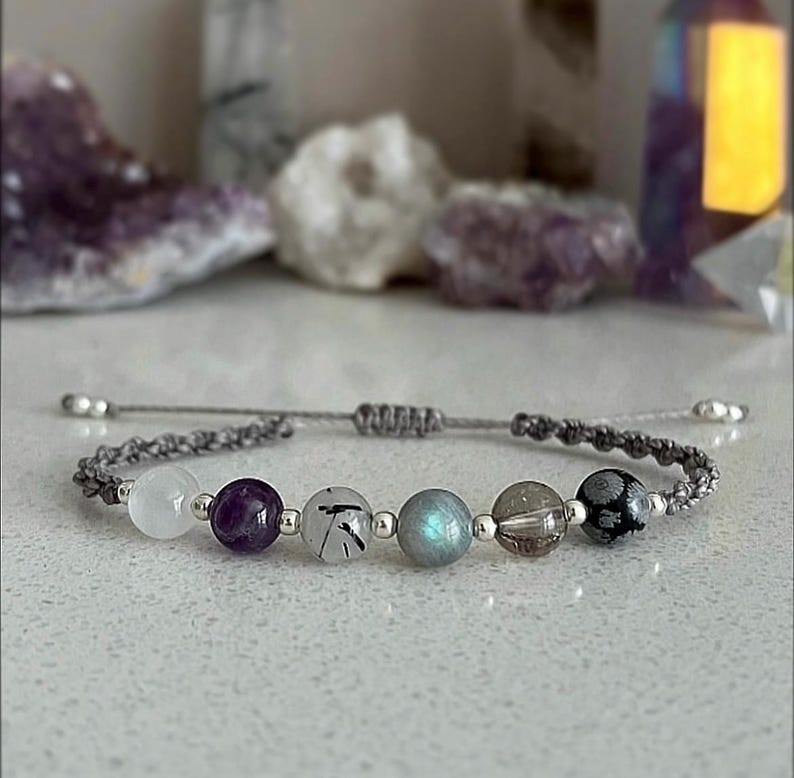 May include: A gray macrame bracelet featuring a series of gemstone beads in white, purple, black, and blue-green hues. The bracelet has silver-colored end beads and a sliding knot closure. The background includes blurred crystals.