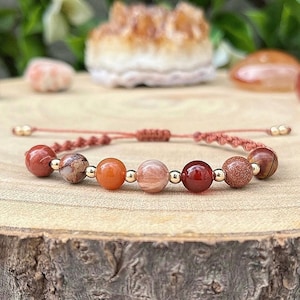 以下が含まれることがあります： 赤、オレンジ、茶色のビーズとゴールドスペーサーが付いたブレスレット。ブレスレットは天然石で作られており、赤い紐で結ばれています。