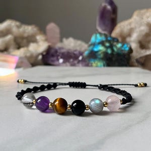 Könnte beinhalten: Ein schwarzes Armband aus Schnur mit sieben Perlen, darunter klare, violette, braune, schwarze, graue und rosa Perlen. Das Armband hat goldene Akzente.